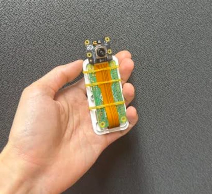 手のひらに載せた本機の試作機。白い専用ケースに小型コンピュータの基板が収まり、オレンジ色のリボンケーブルで上部のカメラモジュールとつながっている。輪ゴムで固定されている。